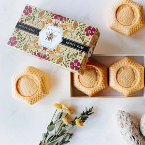 A floral box labeled “BAUDELAIRE - PURE HONEY SOAP 3.5 OZ, 2 BAR GIFT BOX” sits beside two honeycomb-shaped BAUDELAIRE Pure Honey Soaps featuring a bee design—each a gentle, triple-milled soap ideal for sensitive skin—accompanied by a white tray, yellow flowers, and stones on a white surface.