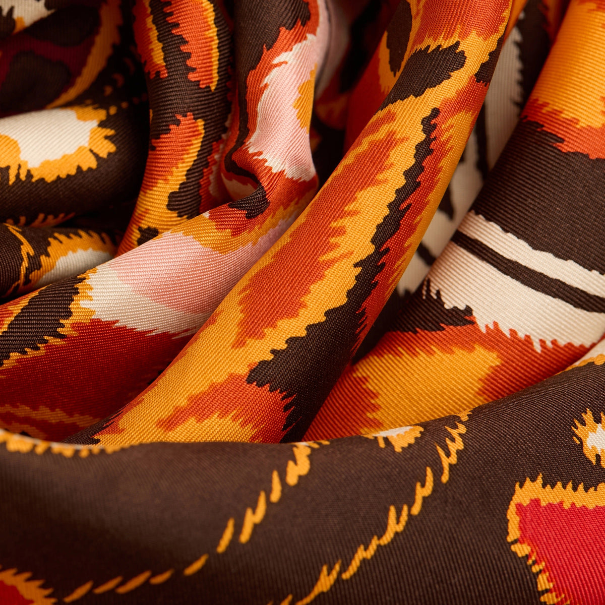 A person in a dark grey knit sweater wears an INOUI EDITIONS PAPILLONS Orange Silk Scarf, featuring bold abstract butterfly designs and “INOUI EDITIONS” on the border, neatly tied around their neck.