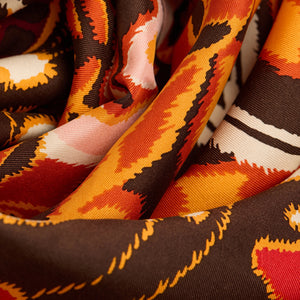 A person in a dark grey knit sweater wears an INOUI EDITIONS PAPILLONS Orange Silk Scarf, featuring bold abstract butterfly designs and “INOUI EDITIONS” on the border, neatly tied around their neck.