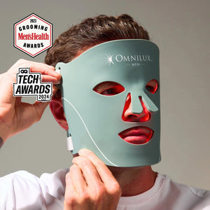 A man intently examines the OMNILUX - MENS MASK, a glowing red LED mask designed for skin rejuvenation with advanced red light therapy, featuring eye, nose, and mouth cutouts.