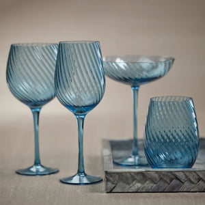 The SAVOY OPTIC SWIRL RED WINE GLASS by ZODAX features a turquoise color with a swirling, ribbed pattern. It boasts a wide bowl, tall stem, and round base, along with a reflective glassy finish. This glass has a capacity of 600 ml.