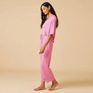 A woman stands barefoot facing away, wearing the SOFTIES Dream Relaxed V-Neck with Capri Lounge Set in Fuschia Stripe—soft jersey pink and white striped pajamas with a loose top and wide-leg pants—against a plain beige background.