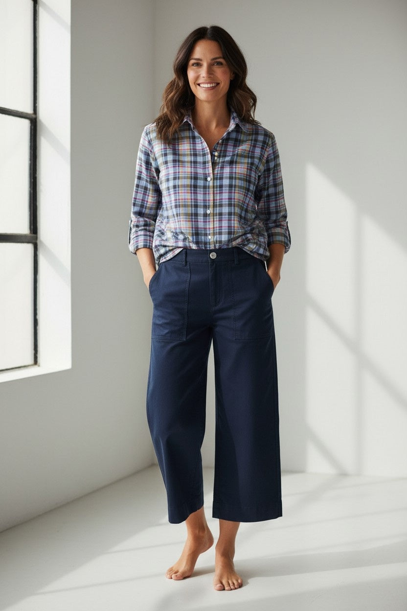 A woman stands barefoot in a sunlit room, smiling with her hands in her pockets, wearing a plaid shirt with rolled sleeves and the FRANK & EILEEN Limerick Wide Leg Carpenter Pant in Classic Navy.