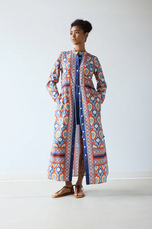 In a minimal room, a person with short curly hair and brown sandals exudes confidence in the HOPE FOR FLOWERS BAND COLLAR SHIRTDRESS IN CHAMBRAY MEDALLION, showcasing modern style from the HOPE FOR FLOWERS brand.