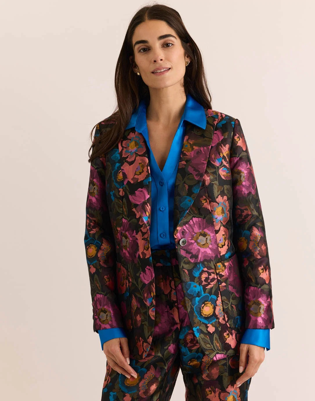A woman with long brown hair is wearing the HINSON WU - CORINNE JACQUARD JACKET IN FLORAL BOUQUET, paired with a bright blue satin shirt underneath, standing against a plain light background.