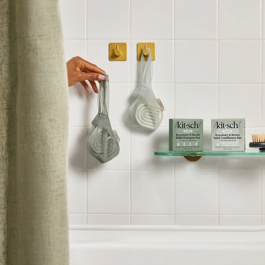 A green, eco-friendly mesh bag from KITSCH sits beside green packaging labeled “KITSCH - ROSEMARY & BIOTIN SHAMPOO BAR SAVER BAG,” designed for storing solid shampoo bars and extending their lifespan. A pink tag reads “Rosemary & Biotin Shampoo Bar.” Both the bag and box are displayed against a white background.