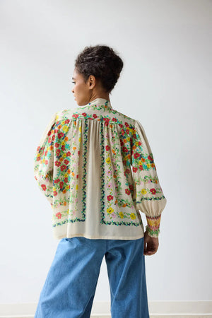 A woman with curly hair looks over her shoulder, wearing the HOPE FOR FLOWERS Smocked Tie Neck Blouse in Custard Floral—a sustainable, cream-colored top with vibrant florals from HOPE FOR FLOWERS, set against a plain, softly lit background.