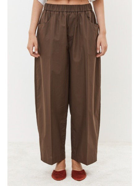 A person is seen from behind wearing MEIMEIJ&#39;s Pull On Cotton Pant, featuring an elastic waistband for comfort. The loose-fitting brown pants with large back pockets are paired with a white top peeking out at the waist, standing on a light surface.