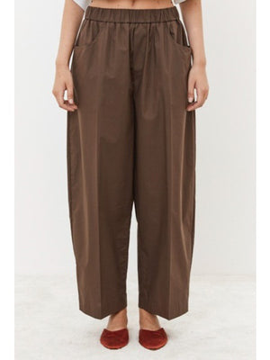 A person is seen from behind wearing MEIMEIJ's Pull On Cotton Pant, featuring an elastic waistband for comfort. The loose-fitting brown pants with large back pockets are paired with a white top peeking out at the waist, standing on a light surface.