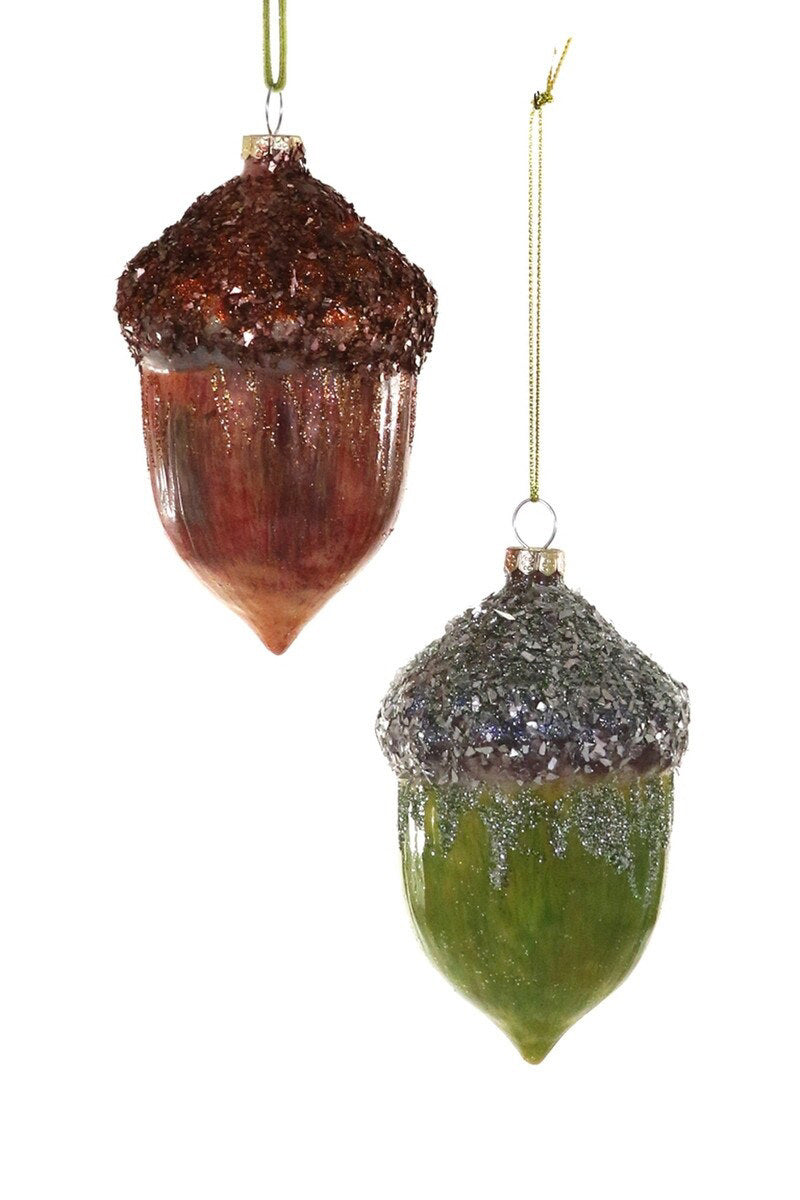 The OLDE OAK ACORN ORNAMENT by CODY FOSTER features two 2.5-inch glass acorn ornaments, one brown and one green with glittery caps, hanging from gold strings against a white background.