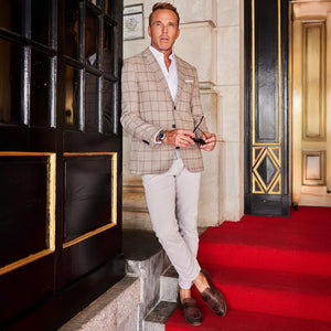A man in sunglasses adjusts his COLLARS AND CO - MAVERICK TRAVEL BLAZER IN BEIGE WINDOWPANE, standing confidently on a red carpet before ornate doors.