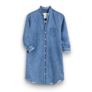 A woman poses, smiling on a stone patio with curly hair flowing freely. She wears the FRANK & EILEEN Mary Ruffle Shirtdress in Vintage Stone Washed Indigo, set against ornate doors and a wall-mounted lamp offering an elegant backdrop.