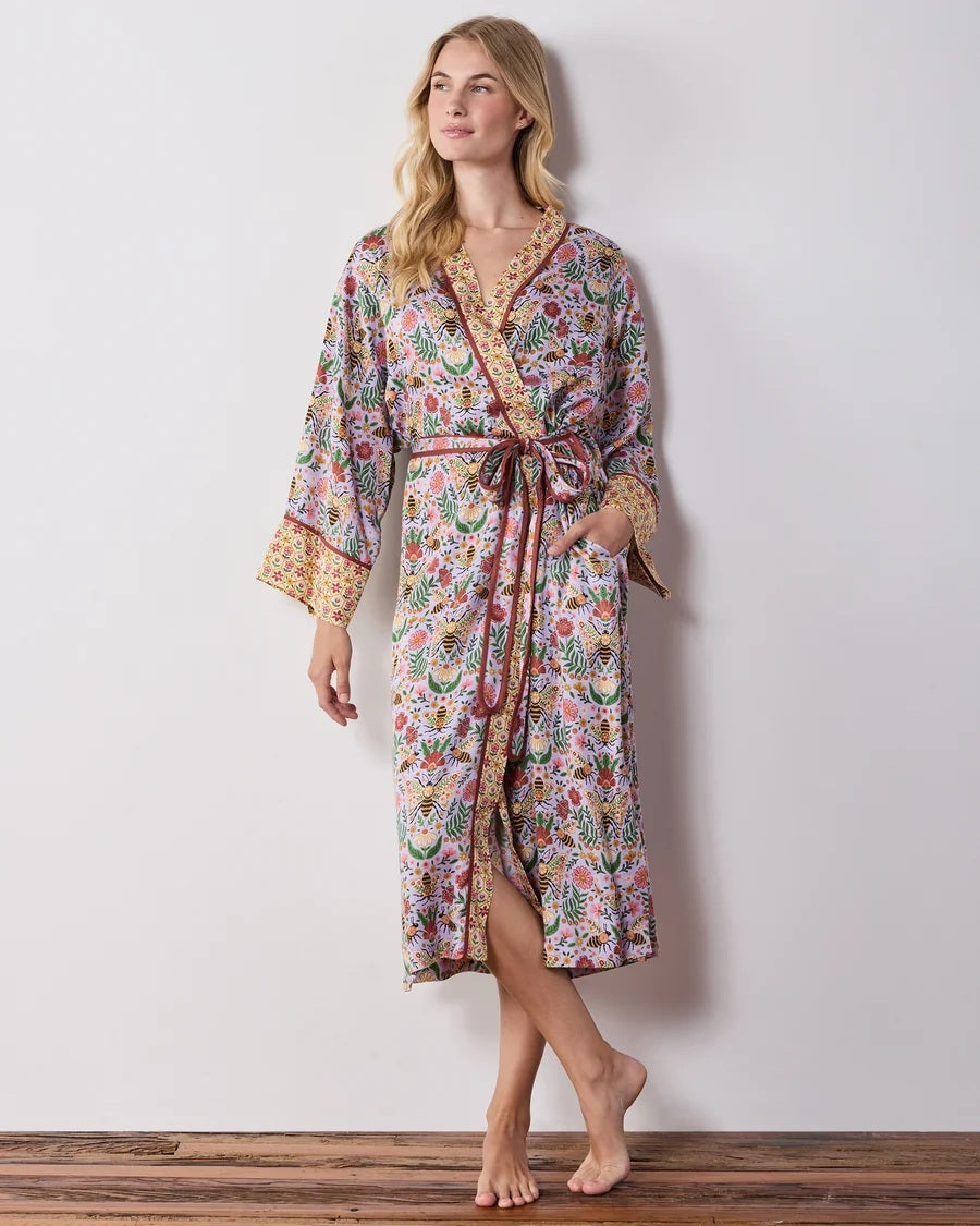 A woman wears a PRINTFRESH - BEE TAPESTRY ECO SATIN ROBE IN LAVENDER, featuring a lavender floral-print with a repeating pattern of bees, flowers, and vines with a contrasting yellow and red patterned trim.