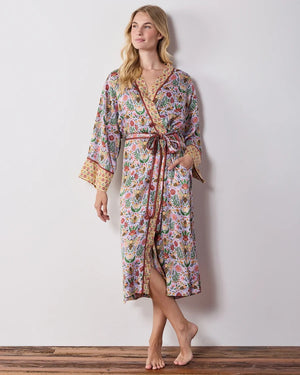 A woman wears a PRINTFRESH - BEE TAPESTRY ECO SATIN ROBE IN LAVENDER, featuring a lavender floral-print with a repeating pattern of bees, flowers, and vines with a contrasting yellow and red patterned trim.
