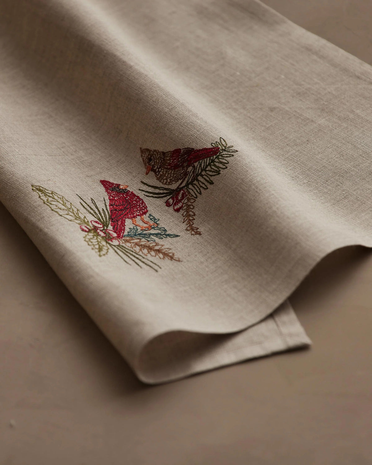 A CORAL & TUSK CHRISTMAS CARDINALS LINEN TEA TOWEL made of embroidered natural flax fiber featuring two cardinals perched on pine branches with berries.