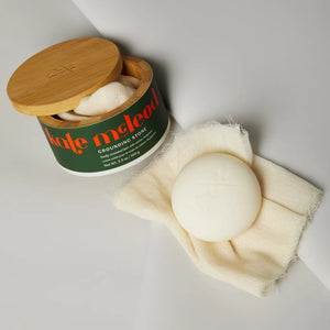 A round white KATE MCLEOD Grounding Stone deep moisturizing lotion bar sits on textured fabric next to an open container with a wooden lid, featuring a dark green label with orange and white text, highlighting its luxurious blend of cocoa butter concentrate and natural essential oils.