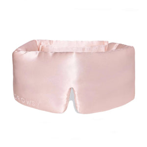 An older person with long gray hair wears a luxurious beige DROWSY - SATIN SLEEP MASK by DROWSY SLEEP CO, made of mulberry silk. In a matching satin top, they stand against a plain white background, ready for beauty sleep.