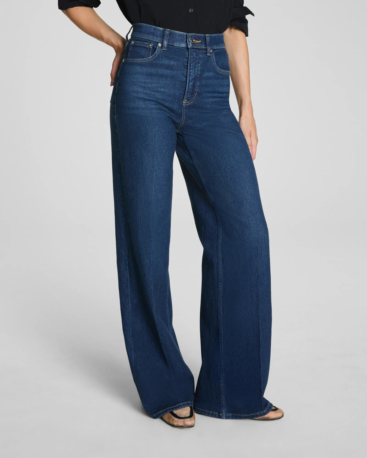 A person with long, dark hair poses against a plain background, wearing a black button-up shirt with rolled sleeves and the SPANX SHAPE AUTHENTIC 360 WIDE LEG JEAN by SPANX. The high-waisted jeans feature wide legs and are crafted from soft 4-way stretch denim, while one hand is tucked casually into a pocket.