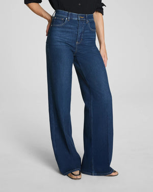 A person with long, dark hair poses against a plain background, wearing a black button-up shirt with rolled sleeves and the SPANX SHAPE AUTHENTIC 360 WIDE LEG JEAN by SPANX. The high-waisted jeans feature wide legs and are crafted from soft 4-way stretch denim, while one hand is tucked casually into a pocket.