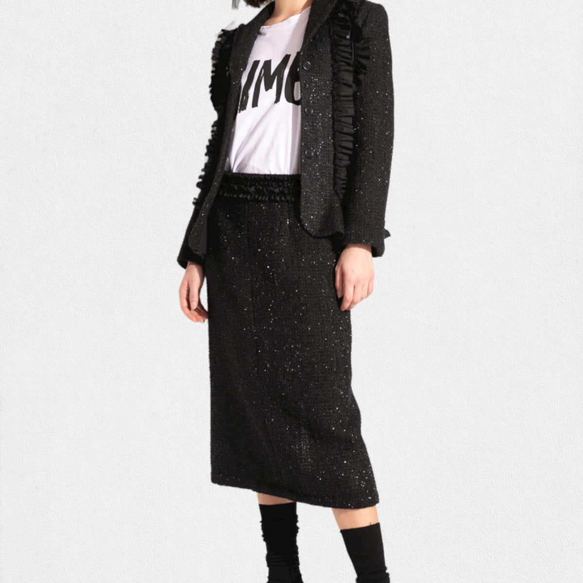 A person is wearing a stylish, textured black MEIMEIJ Lurex Tweed suit, featuring a tailored jacket with fringe details and the MEIMEIJ - LUREX TWEED SKIRT. Underneath the jacket is a white T-shirt with bold black text. The outfit is paired with black socks and black shoes.