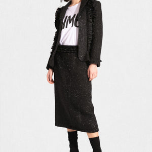A person is wearing a stylish, textured black MEIMEIJ Lurex Tweed suit, featuring a tailored jacket with fringe details and the MEIMEIJ - LUREX TWEED SKIRT. Underneath the jacket is a white T-shirt with bold black text. The outfit is paired with black socks and black shoes.