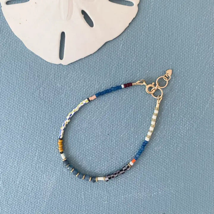 The ALICE RISE - SAPPHIRE GEMSTONE BRACELET by ALICE RISE, featuring multicolored beads and gold clasps, rests on a blue textured surface beside a white sand dollar—a striking example of elegant jewelry.