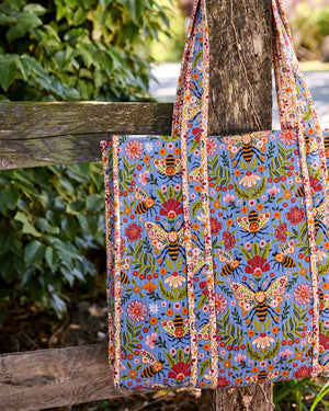 The PRINTFRESH - Bee Tapestry Traveler Tote in Delphinium Blue by PRINTFRESH, featuring vibrant floral, butterfly, and bee designs on organic cotton, hangs on a rustic wooden fence outdoors with lush green foliage in the background.