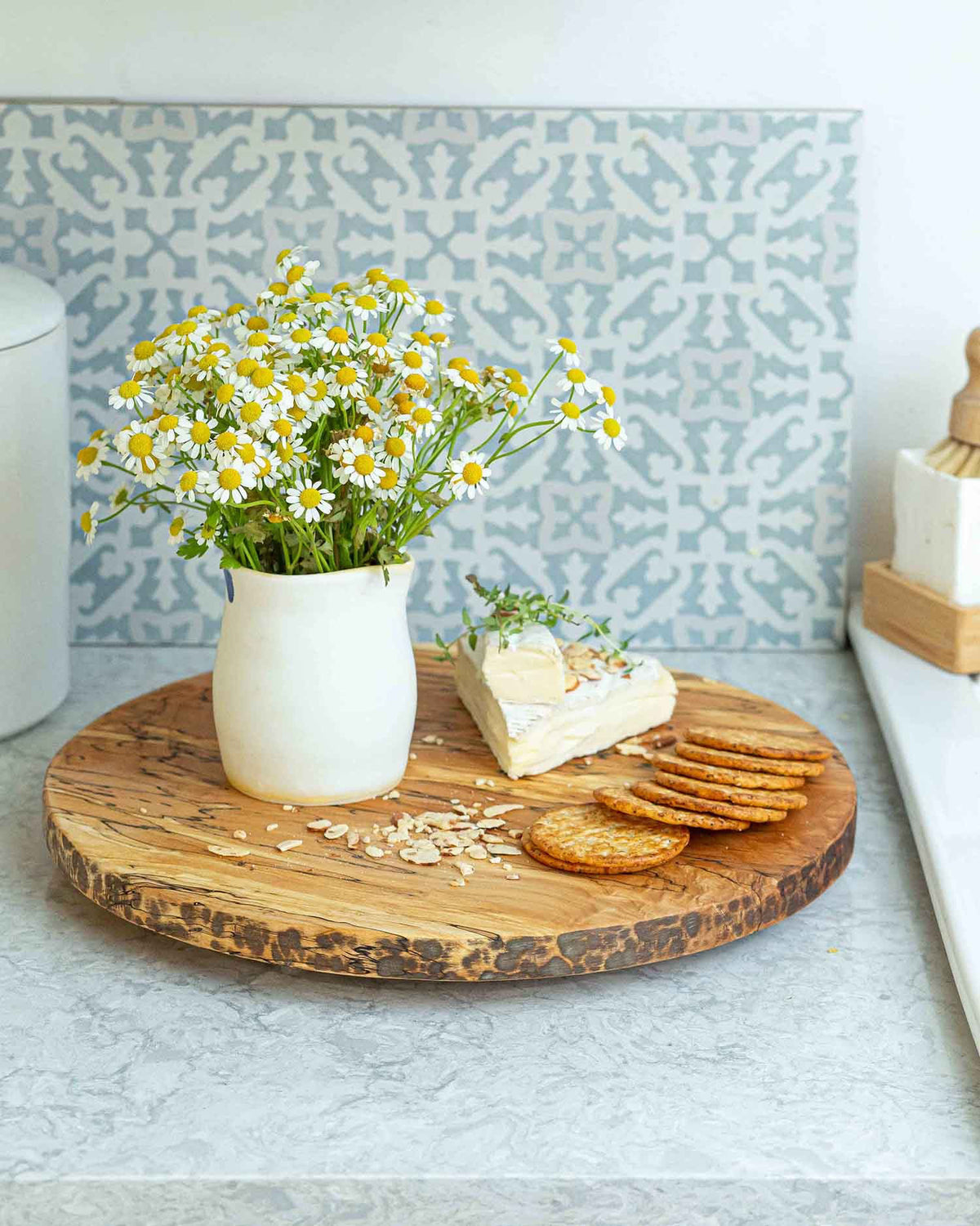 A small white vase filled with yellow and white daisy flowers sits on a PETERMANS BOARDS &amp; BOWLS INC SPENCER PETERMAN - SPALTED LAZY SUSAN 20&quot; along with a wedge of soft cheese topped with herbs and a few round crackers. The spinning tray rests on a kitchen counter, showcasing a patterned tile backsplash in the background.