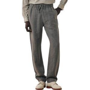 A man stands against a plain background wearing a light gray long-sleeve henley shirt, HARTFORD - LINEN JOGGY PANTS IN ARMY, and light-colored shoes, with his hands in his pockets.