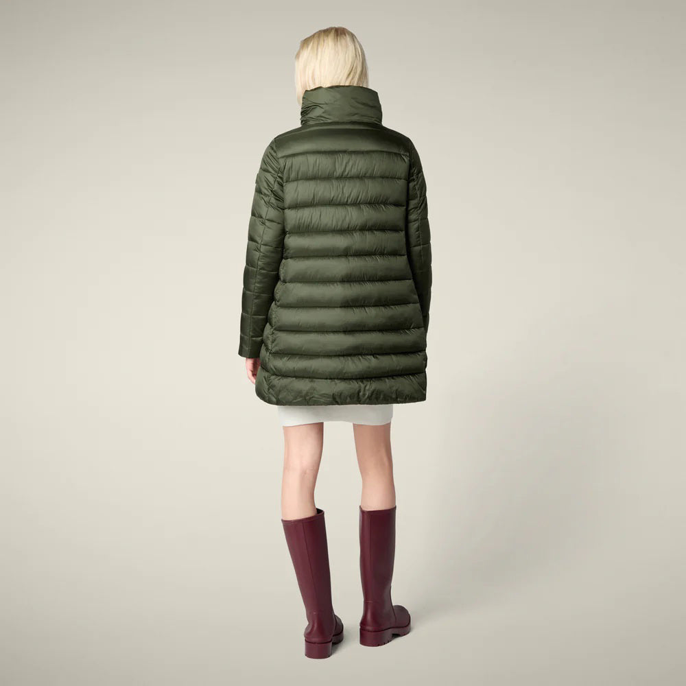 A woman in the SAVE THE DUCK - LYDIA PUFFER COAT by SAVE THE DUCK, paired with a white dress, maroon knee-high boots, and holding a fluffy light pink clutch, stands against a plain beige background.