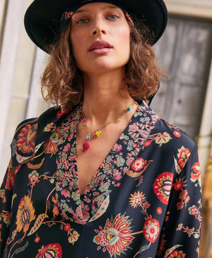 The MOMONI - FELICIEN DRESS by MOMONI is a long-sleeved, ankle-length black crepe de chine dress with a vibrant floral and paisley print in red, orange, and green, finished with patterned borders at the hem and cuffs.