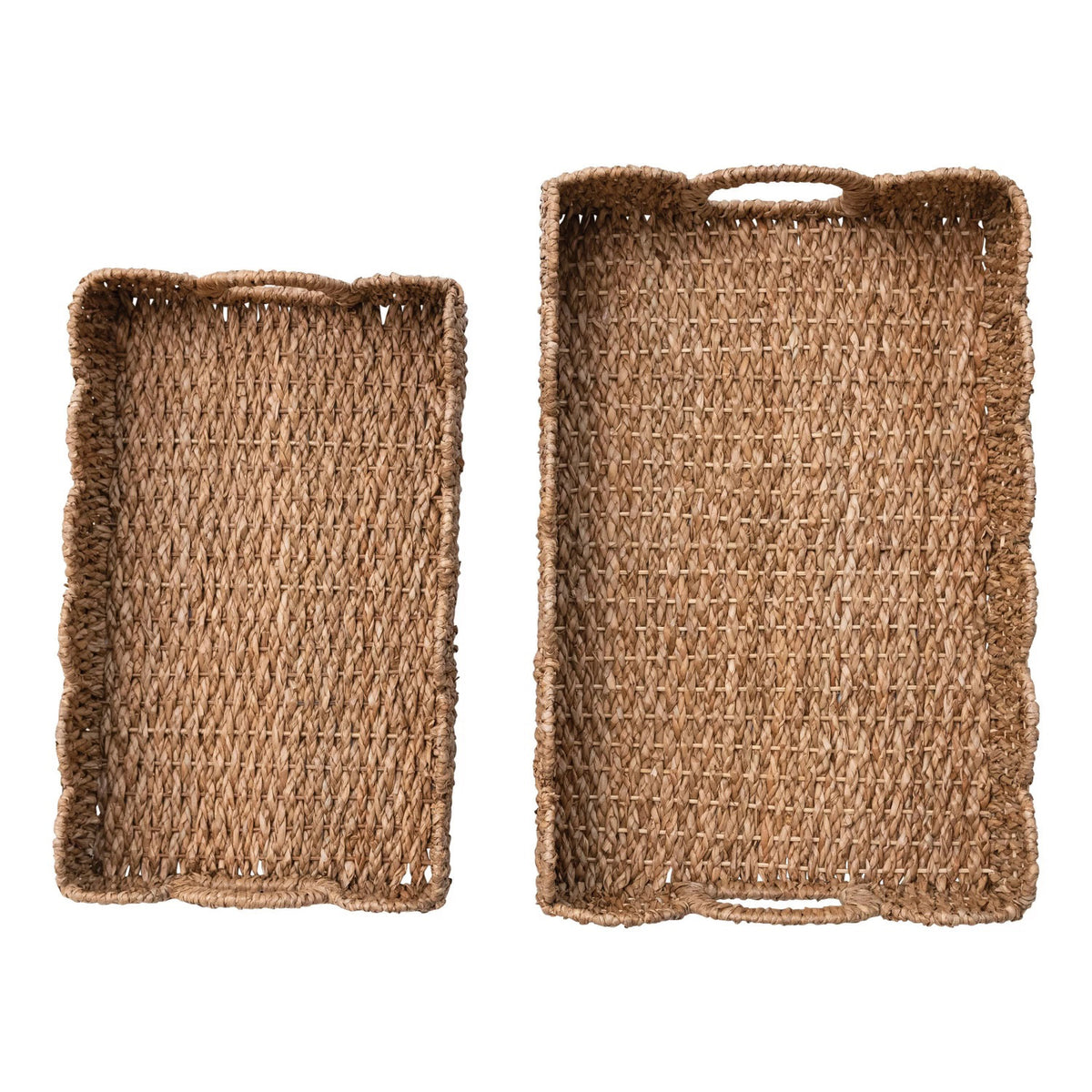 Two rectangular woven baskets from Creative Co-op are placed vertically on a white surface. The top basket, a BRAIDED BANKUAN TRAY WITH HANDLE (SMALL), contains a cup and saucer, a bowl with cereal, and some papers. The bottom basket is empty and has a slightly scalloped edge.