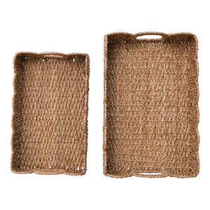 Two rectangular woven baskets from Creative Co-op are placed vertically on a white surface. The top basket, a BRAIDED BANKUAN TRAY WITH HANDLE (SMALL), contains a cup and saucer, a bowl with cereal, and some papers. The bottom basket is empty and has a slightly scalloped edge.