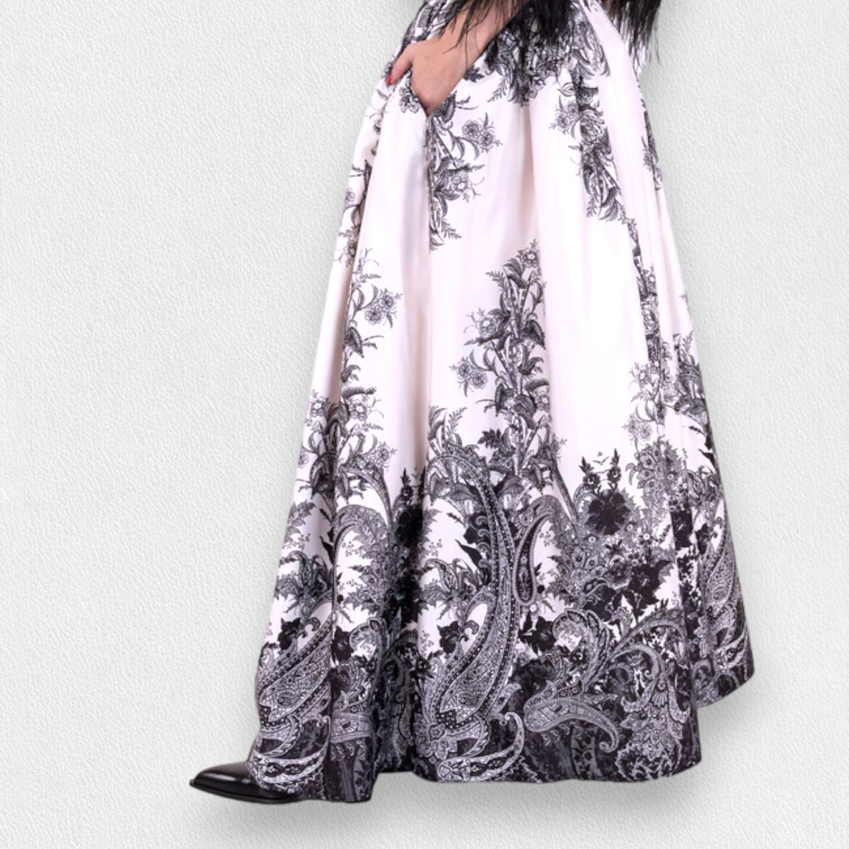 A woman stands against a plain gray backdrop wearing a sheer black blouse with ruffle details at the chest and MILVA&#39;s LONG MAXI SKIRT, featuring a detailed black-and-white pattern. The high-waisted, floor-length skirt, with welt pockets and a side closure with concealed zip, cinches at her gathered waist. She wears black shoes.