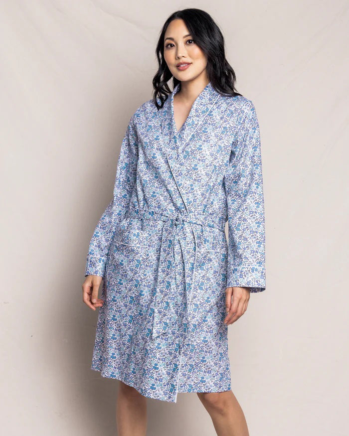 The PETITE PLUME - FLEUR D&#39;AZURE ROBE by PETITE PLUME is a knee-length, long-sleeved bathrobe adorned with a vintage-inspired floral print in blue and white hues. Crafted from the finest quality cotton, the robe features a shawl collar and includes a matching fabric belt tied at the waist. It offers a relaxed fit ideal for comfortable lounging in luxurious sleepwear.