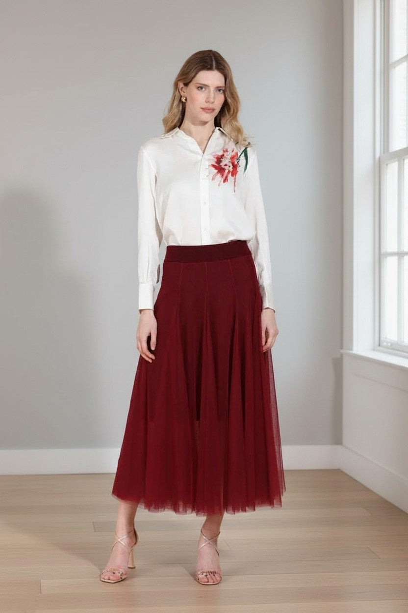 A woman stands indoors wearing a white blouse with floral embroidery paired with the JESSIE LIU Tulle Midi Skirt with Merino Wool Lining in Wine Red. She has wavy hair, neutral makeup, and strappy nude heels as light streams in from windows on the right.