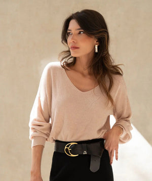 A woman with wavy brown hair wearing a light beige sweater, large hoop earrings, and a black skirt with a wide black belt featuring a gold buckle, stands against a plain, softly lit background.