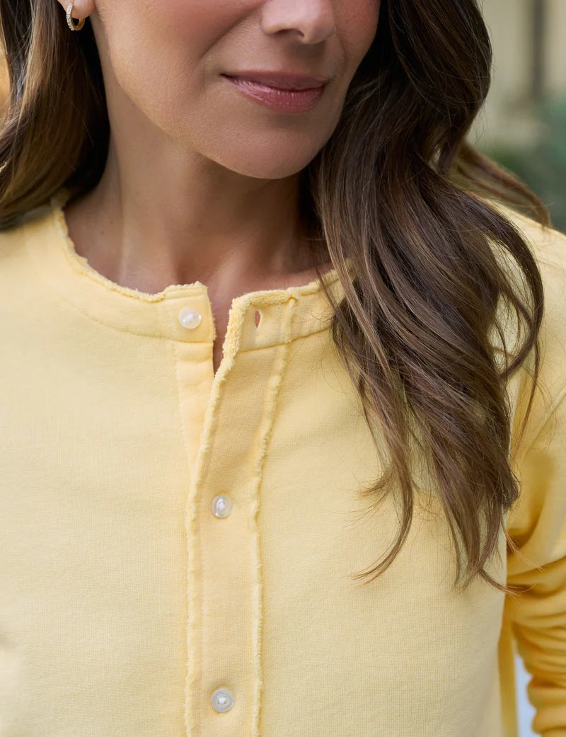 A woman with long brown hair stands outdoors, her back turned, wearing the FRANK &amp; EILEEN Jackie Sweatshirt Cardigan in Lemon. She is on a sunny patio surrounded by plants and large pottery.
