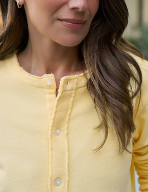 A woman with long brown hair stands outdoors, her back turned, wearing the FRANK & EILEEN Jackie Sweatshirt Cardigan in Lemon. She is on a sunny patio surrounded by plants and large pottery.