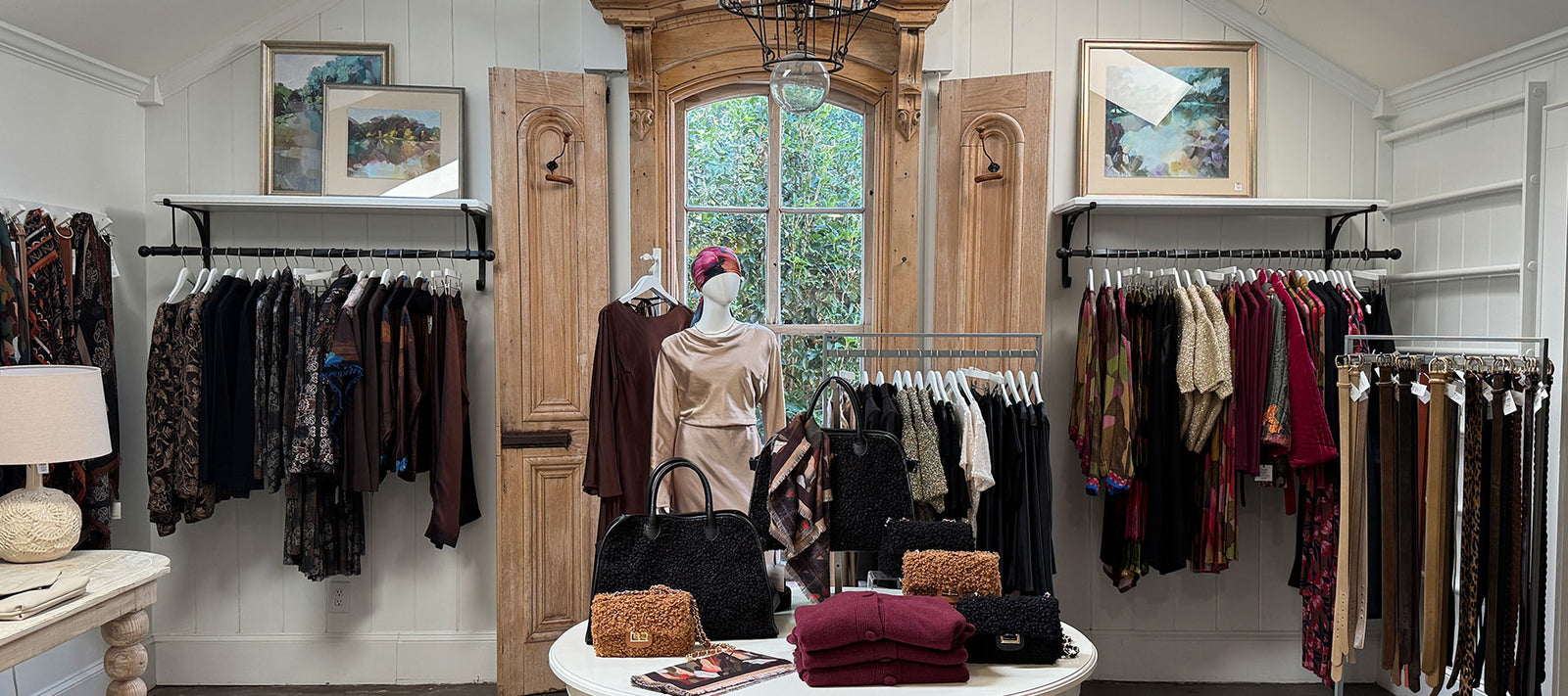 A boutique clothing store interior with racks of clothes, a mannequin in a pink dress, handbags and folded sweaters on a round table, and framed art on the walls. Large wooden window at the center.