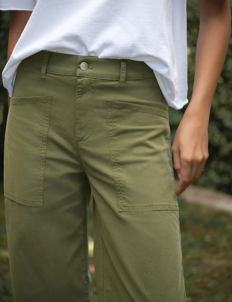 Outdoors on grass, a person wears the FRANK &amp; EILEEN Bray Everyday Barrel Pant in Italian Peached Twill Olive with large front pockets and a white top. One hand is in a pocket, with greenery blurred in the background.