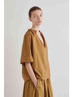 A person wearing the MEIMEIJ Gathered Neck Blouse in Off White by MEIMEIJ stands against a plain light background, looking calmly at the camera with hands in pockets.