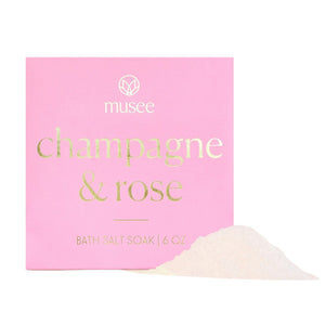 A pink box labeled “MUSEE - CHAMPAGNE & ROSE MINI BATH SALT SOAK” in gold lettering, with a small pile of white bath salt crystals displayed in front.