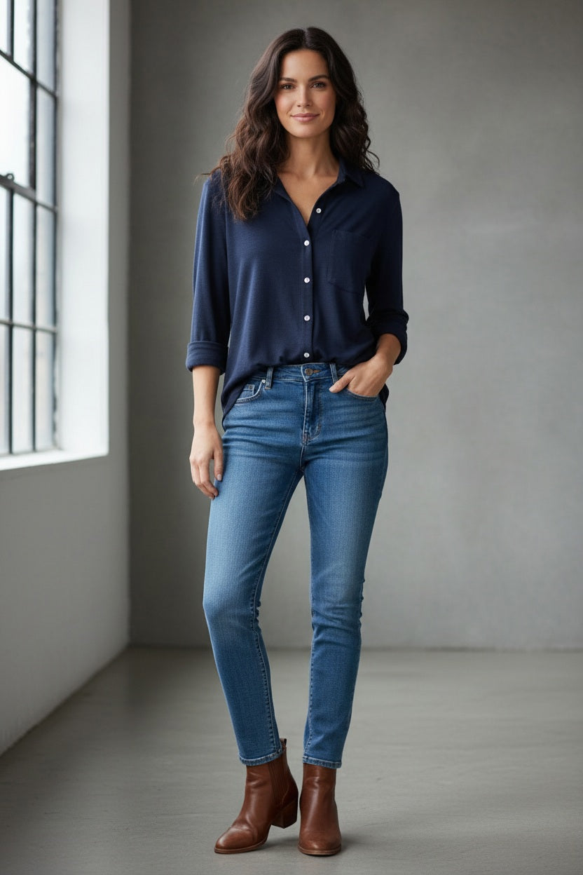 A woman stands in a minimal room, smiling with one hand in her pocket, wearing the FRANK & EILEEN - Waverley Classic Button Up Tee in Vintage Jersey Classic Navy from the California Heritage Collection, paired with blue jeans and brown ankle boots. A large window to the left fills the space with natural light.