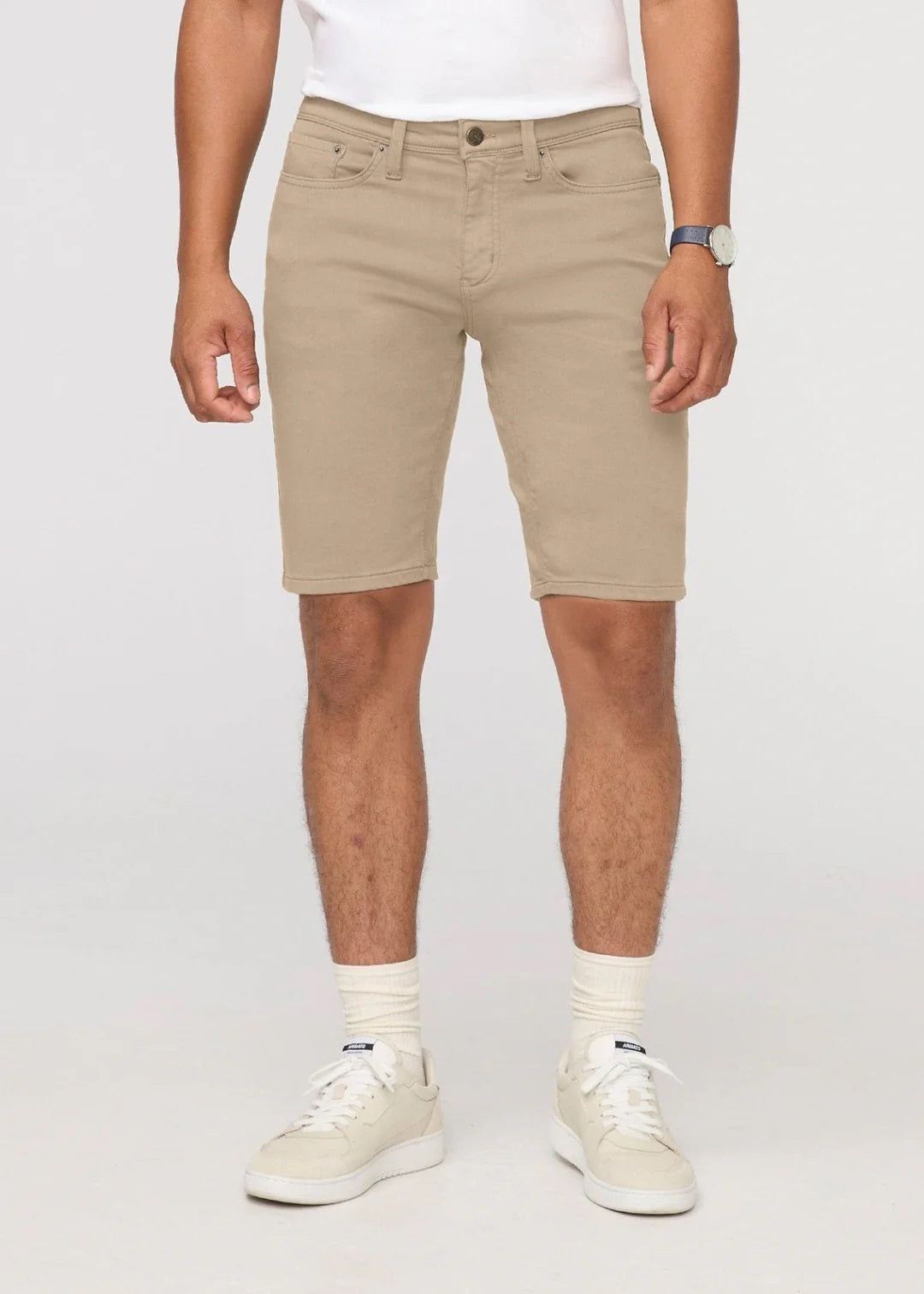 A person wearing a white t-shirt, DUER No Sweat Relaxed Shorts in Desert Khaki, white crew socks, white sneakers, and a wristwatch stands against a plain light background.
