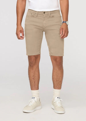 A person wearing a white t-shirt, DUER No Sweat Relaxed Shorts in Desert Khaki, white crew socks, white sneakers, and a wristwatch stands against a plain light background.
