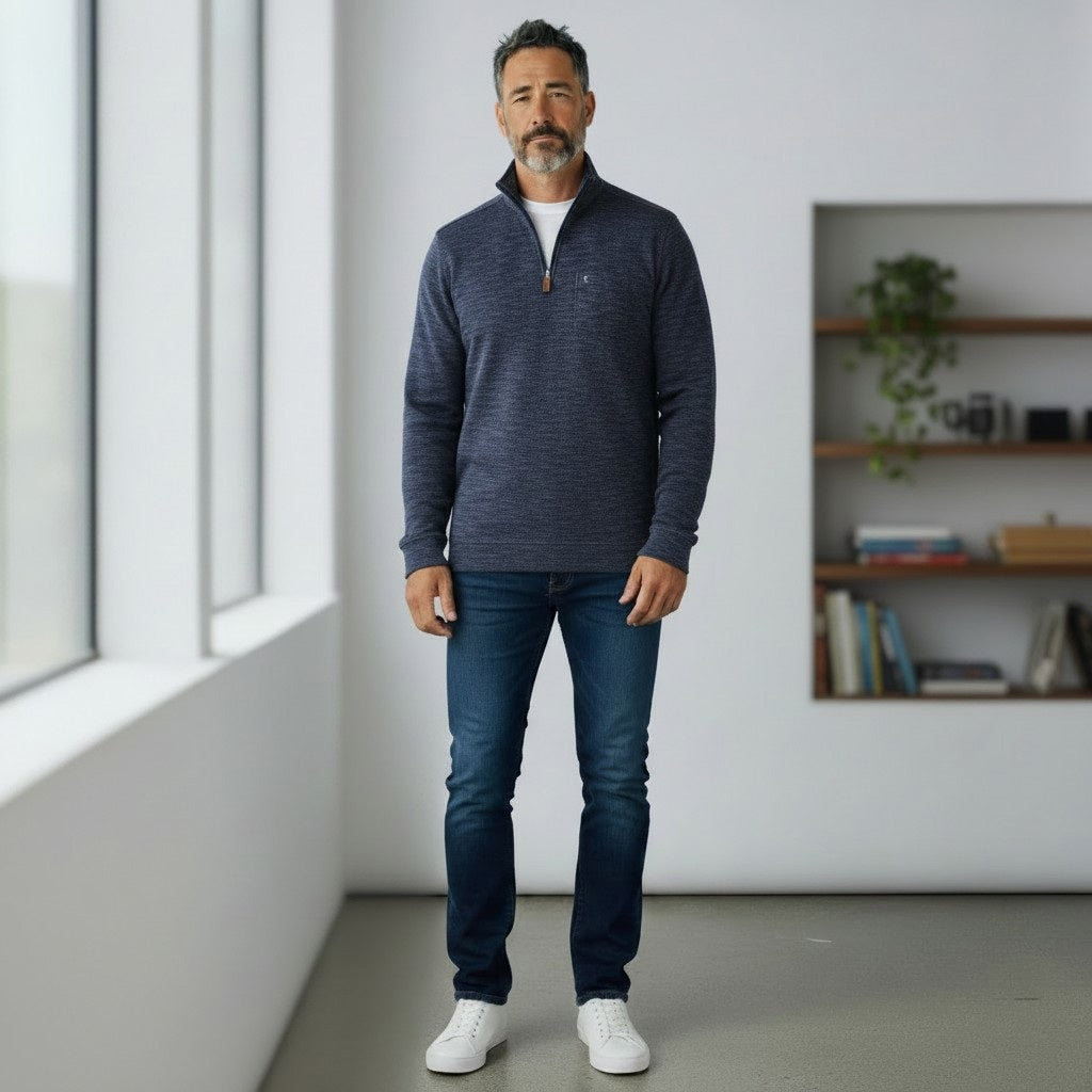 A man with short dark hair and a beard wears the CRIQUET - CROSSROADS Pullover in Moody Blue from CRIQUET over a white shirt and blue jeans. He stands facing forward against a plain white background.