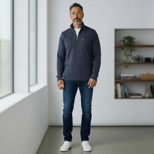 A man with short dark hair and a beard wears the CRIQUET - CROSSROADS Pullover in Moody Blue from CRIQUET over a white shirt and blue jeans. He stands facing forward against a plain white background.
