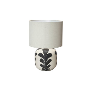 The CREATIVE COOP BLACK AND CREAM STONEWARE TABLE LAMP features a round ceramic base with black leaf designs and a light beige linen shade, set against a plain white background.