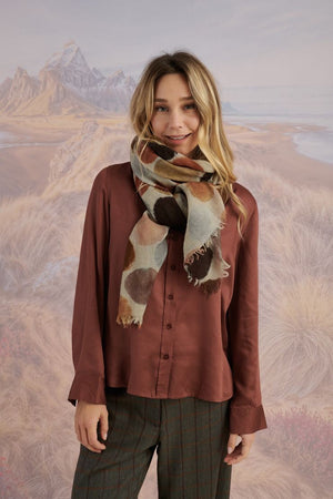 A woman with blonde hair displays the STORIATIPIC - BETTY WOOL SCARF, a large rectangular wool accessory with earth-toned polka dots, set against a scenic mountain landscape.